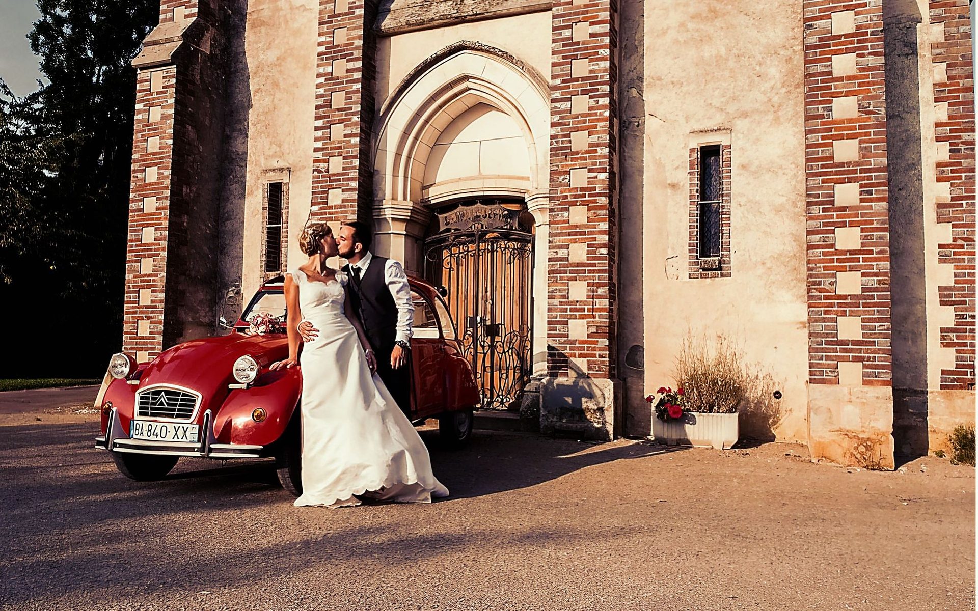 Baiser des mariés devant une 2CV rouge et une chapelle – photographe mariage Île-de-France